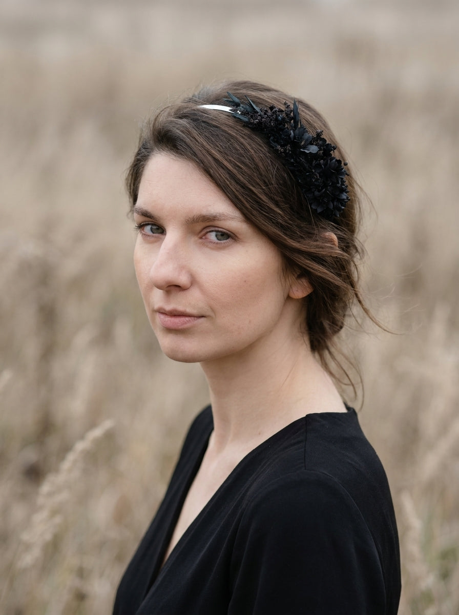 A woman with brown hair wearing a black floral headband and a black top, photographed outdoors against a blurred field of dry grass.