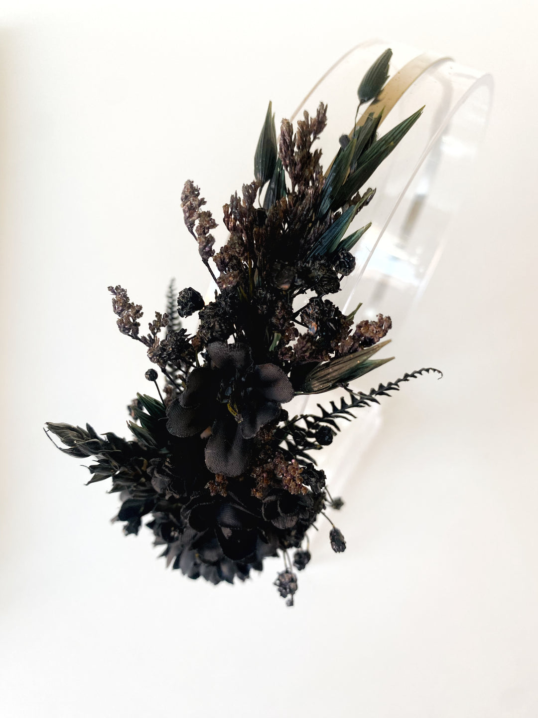 A floral hairpiece or headpiece featuring dark black and brown flowers, green leaves, and small seed heads attached to a clear transparent band, shown against a plain white background.