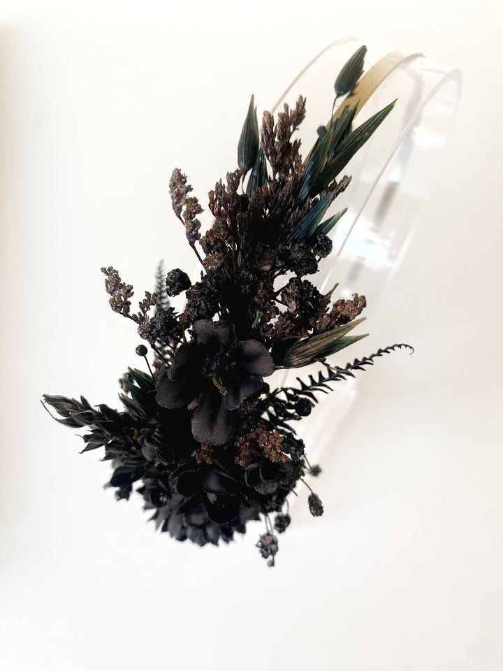A floral hairpiece or headpiece featuring dark black and brown flowers, green leaves, and small seed heads attached to a clear transparent band, shown against a plain white background.