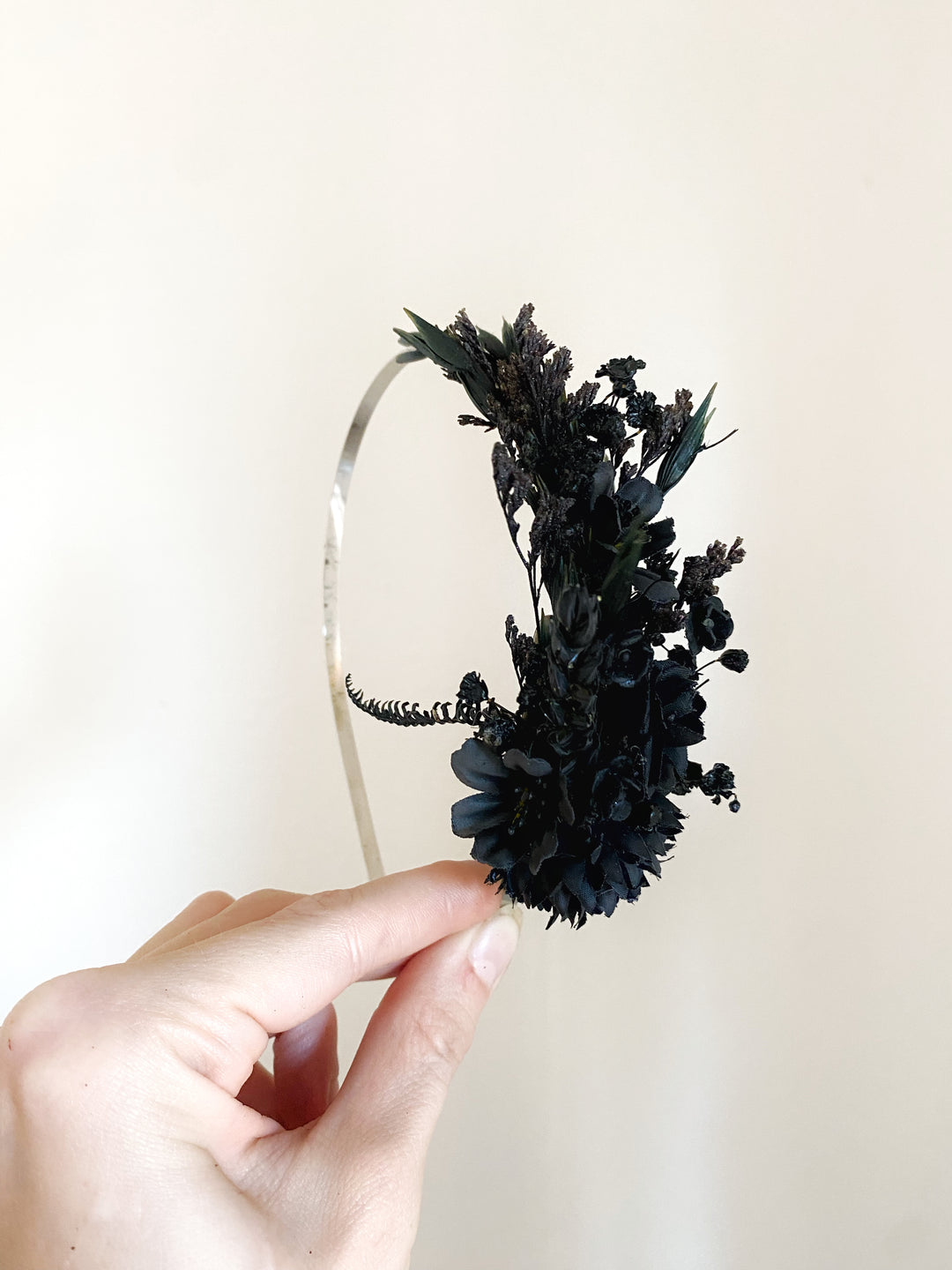 A hand holding a  headband adorned with a large, dense cluster of dried black flowers and small dark green leaves, set against a plain off-white background.