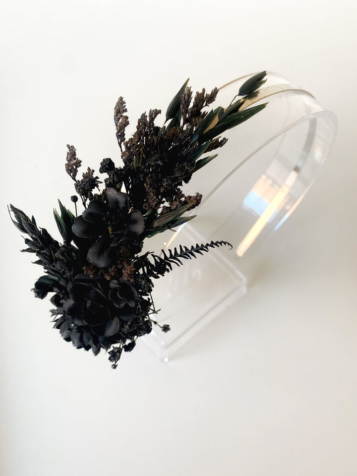 A black flower and foliage hairpiece displayed on a clear acrylic stand against a plain white background.