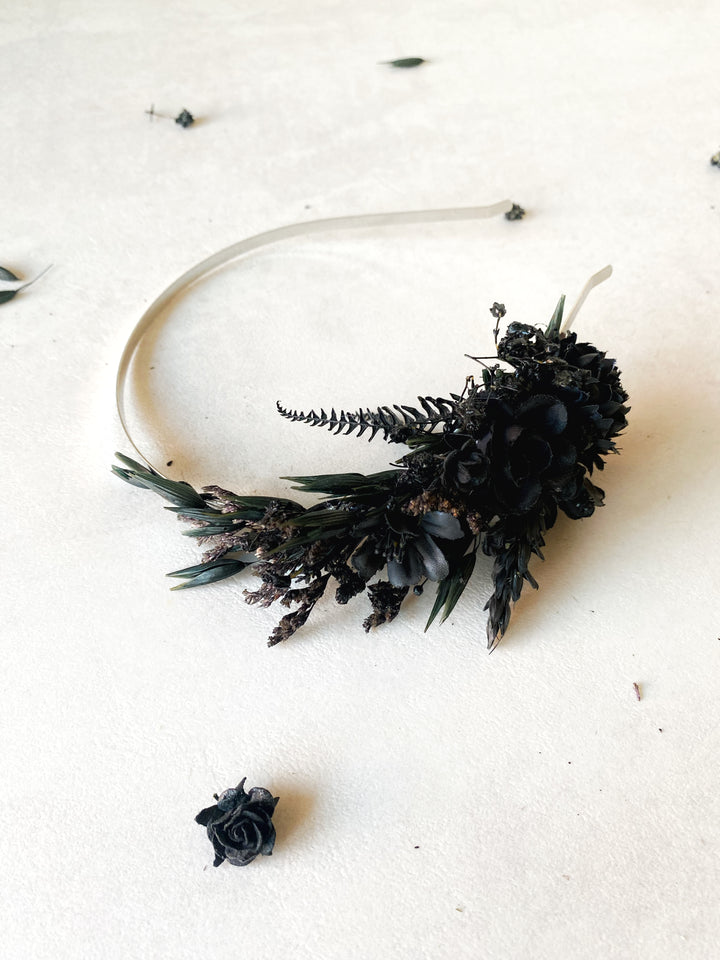 A black floral headband featuring black roses, ferns, and small purple accents on a clear elastic band, displayed on a textured off-white surface.