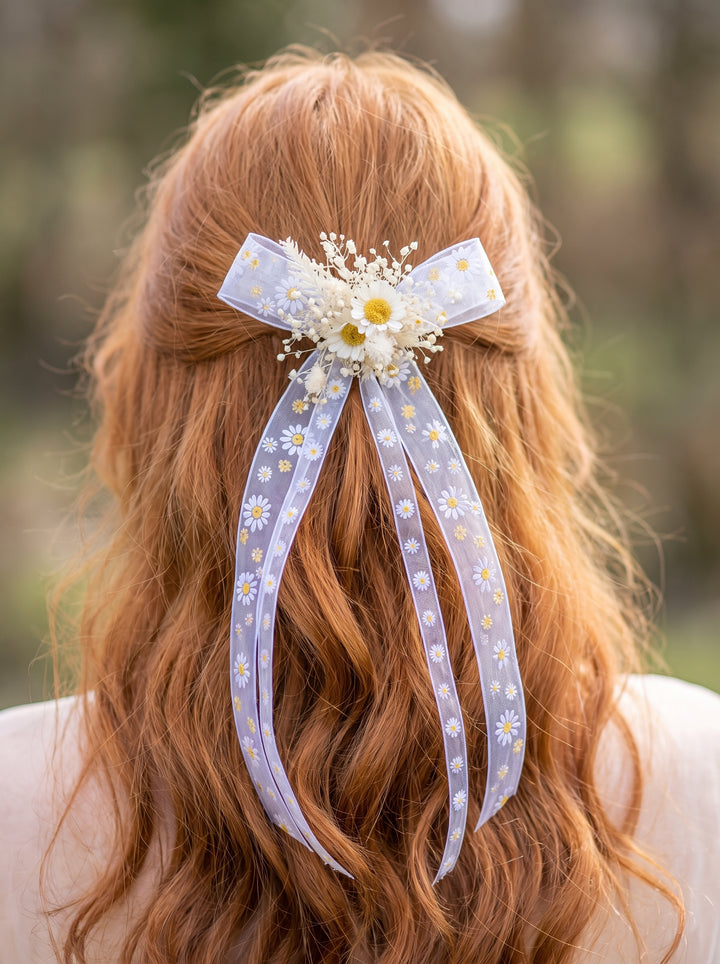 A red-haired woman shown from behind wearing a ribbon hair accessory with white daisy prints, a white bow, and small dried flowers.