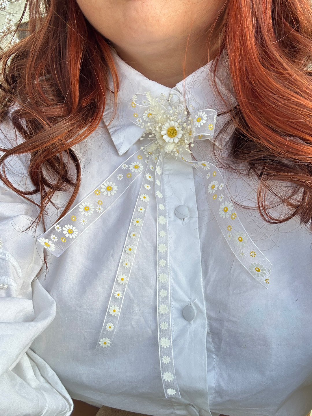 A close-up of a white collared shirt featuring a sheer white ribbon bow decorated with small white daisies and yellow centers, styled with reddish-brown hair.
