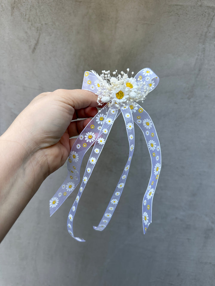 A hand holding a ribbon hair accessory with a daisy pattern and dried white flowers, set against a textured gray background.