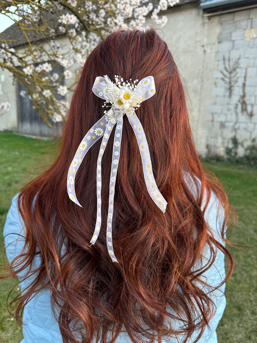 A long, wavy auburn hair accessory with a white and patterned ribbon bow and daisy embellishments, shown clipped to a person's hair outdoors.