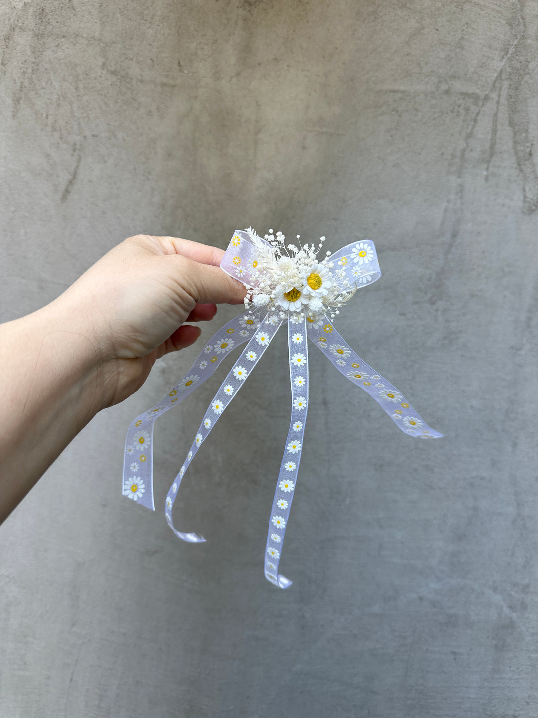 A hand holding a small decorative hair accessory featuring a white and light daisy-patterned ribbon bow with dried white flowers and a yellow center, set against a textured gray background.