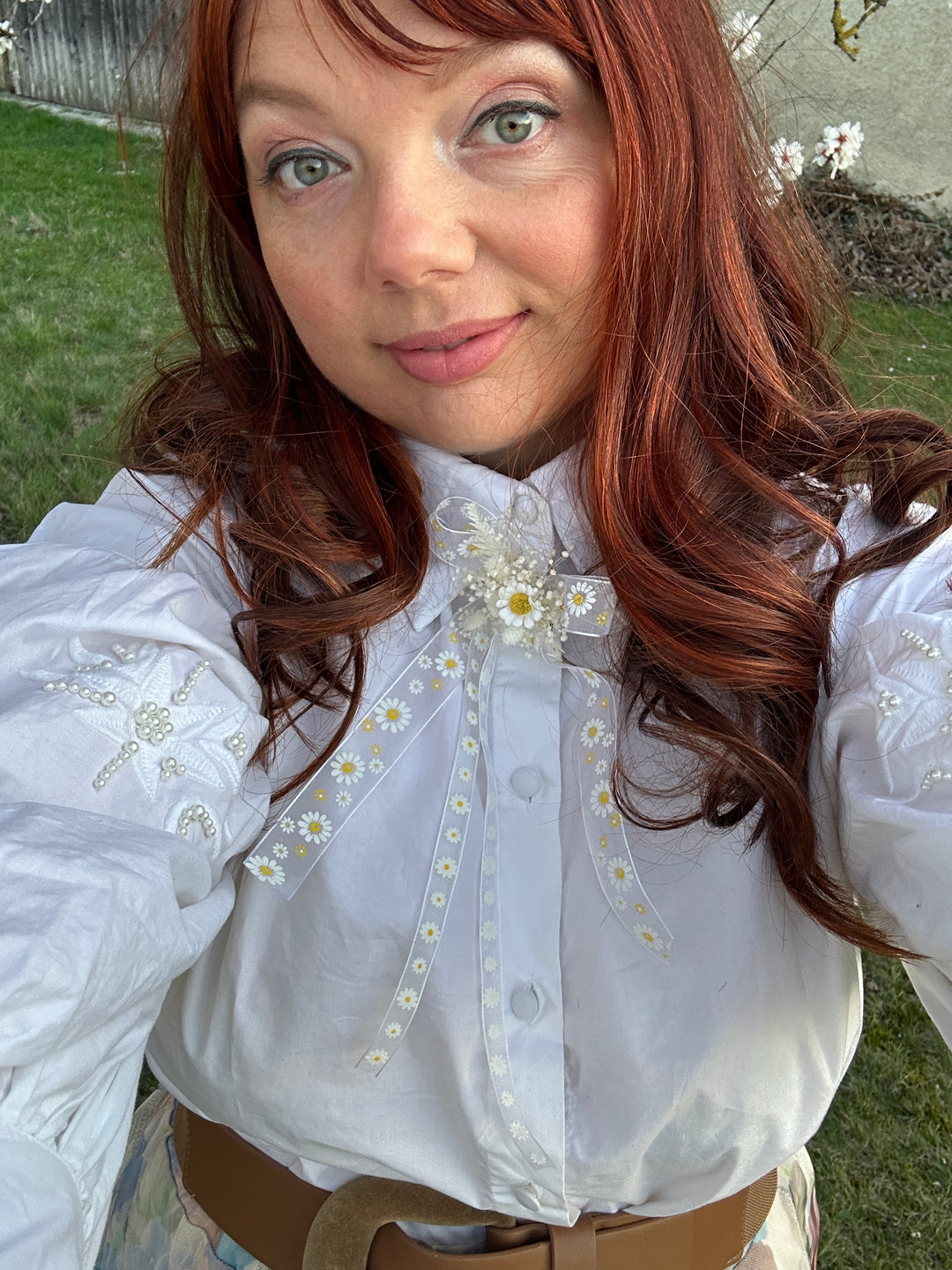 A close-up selfie of a person wearing a white long-sleeve blouse with delicate daisy embroidery and a sheer embroidered chest panel, paired with a brown belt, outdoors on grass.