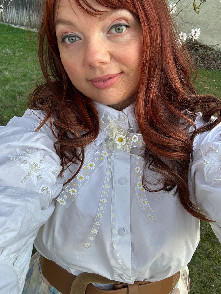 A close-up selfie of a person wearing a white long-sleeve blouse with delicate daisy embroidery and a sheer embroidered chest panel, paired with a brown belt, outdoors on grass.