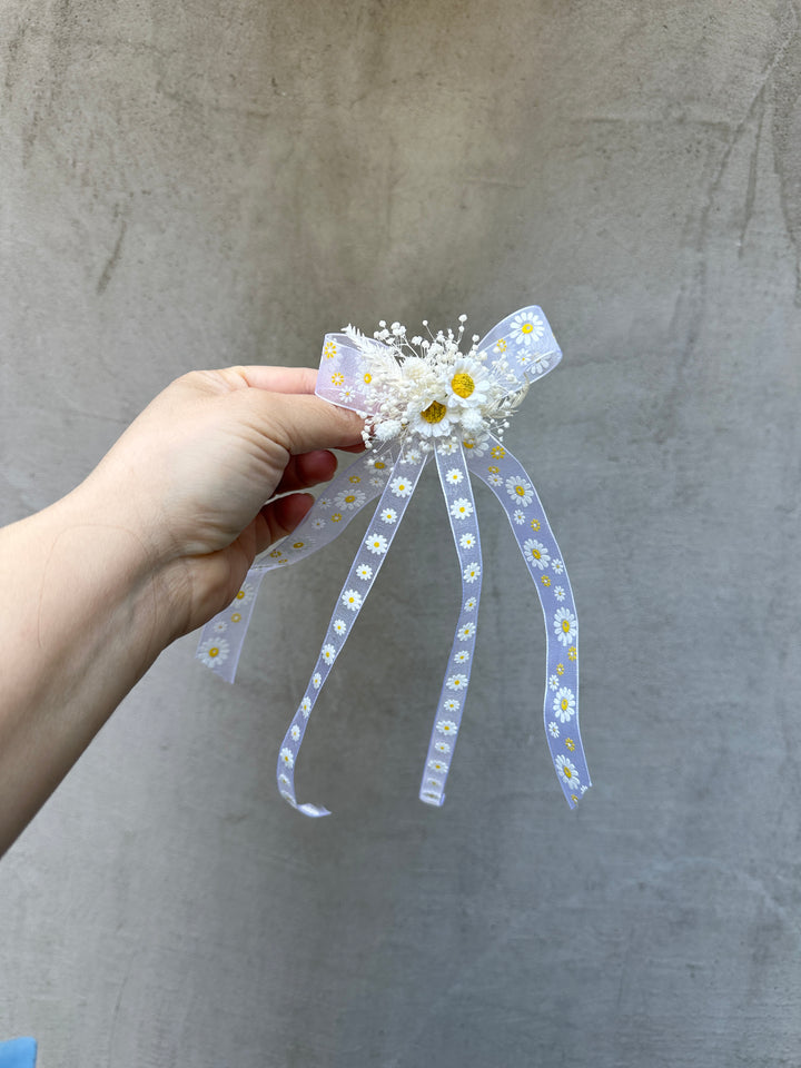 A hand holding a decorative hair accessory featuring a white and daisy patterned ribbon bow with small white flowers and yellow centers, set against a textured gray concrete background.