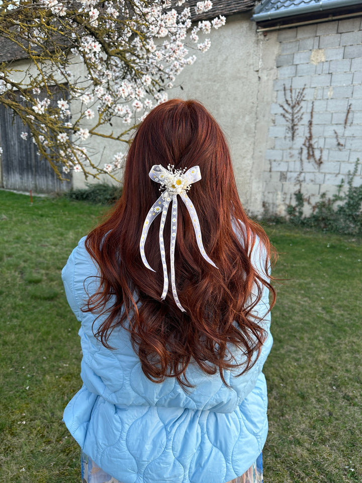 A person outdoors on grass wearing a light blue quilted jacket, viewed from behind. The person has long wavy reddish-brown hair and is wearing a white ribbon hair accessory with a small yellow flower.