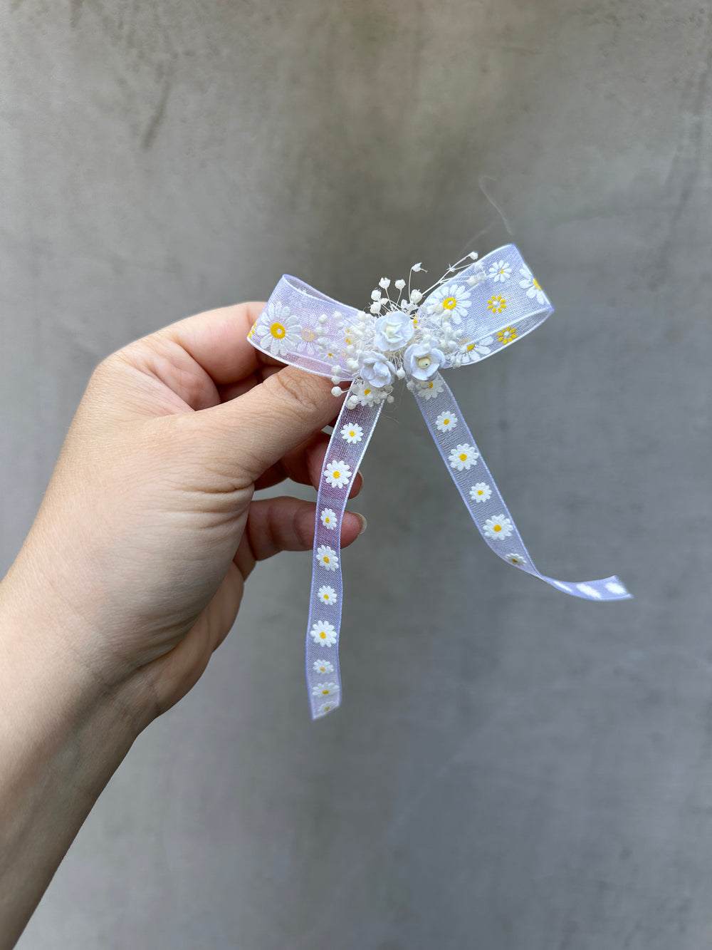 A hand holding a light white ribbon hair bow decorated with white and yellow daisy flowers, set against a neutral gray textured background.