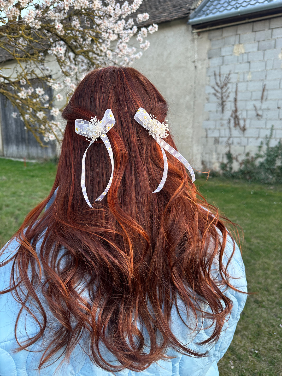 Two white hair clips with clear embellishments and ribbon details, shown in use on a person's auburn hair outdoors.