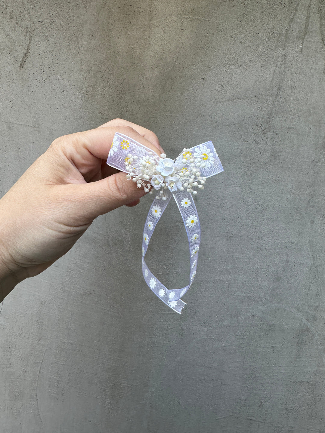 A hand holding a small decorative hair bow made of white faux flowers  on a sheer white ribbon with a white daisy pattern, set against a textured gray concrete background.