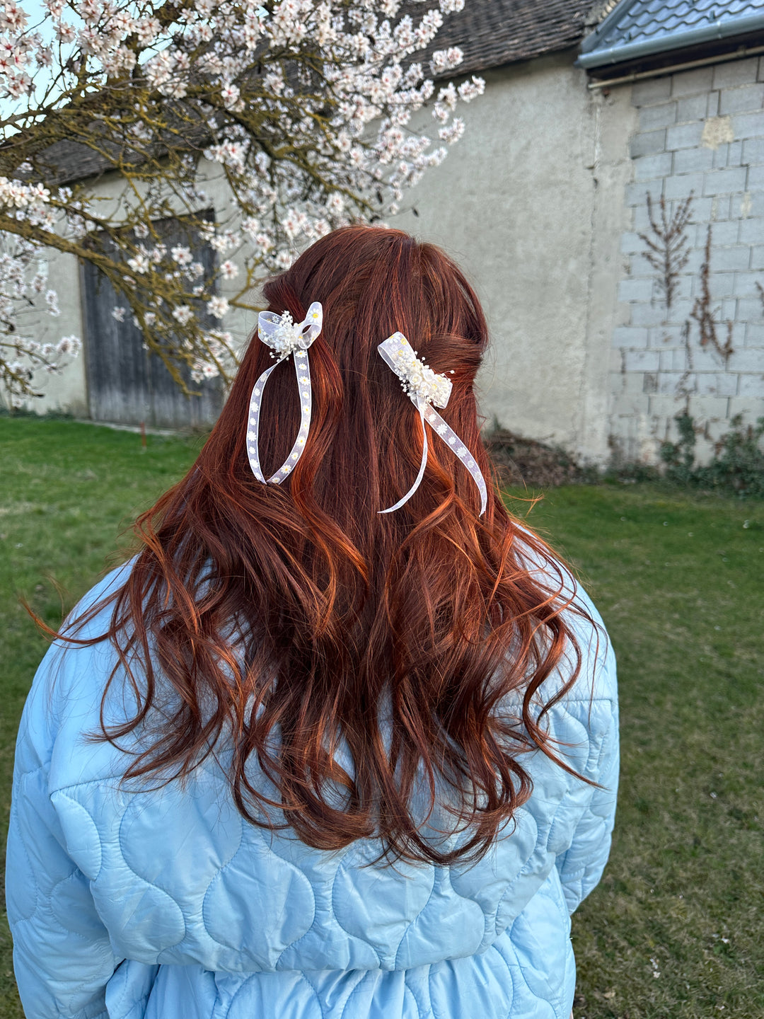A person outdoors with long wavy reddish-brown hair wearing two white lace hair clips, wearing a light blue quilted jacket. A grassy lawn and flowering tree branches are visible in the background.