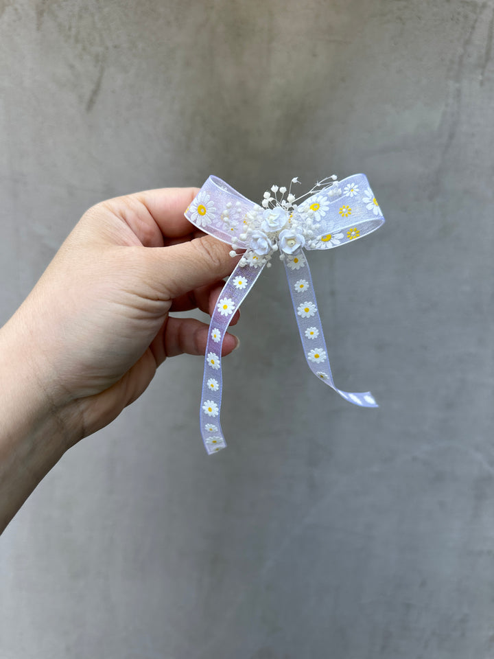A hand holding a white ribbon hair accessory decorated with white daisy patterns, pearl-like beads, and small white flowers, set against a textured gray background.