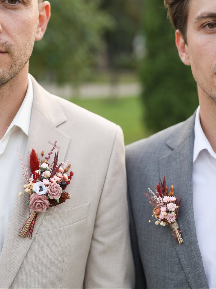 Two men in suits wearing matching floral boutonniere boutonnieres outdoors. The man on the left wears a light beige suit and the man on the right wears a gray suit, both with white shirts and matching floral boutonnieres featuring dusty pink, burgundy, and cream elements.