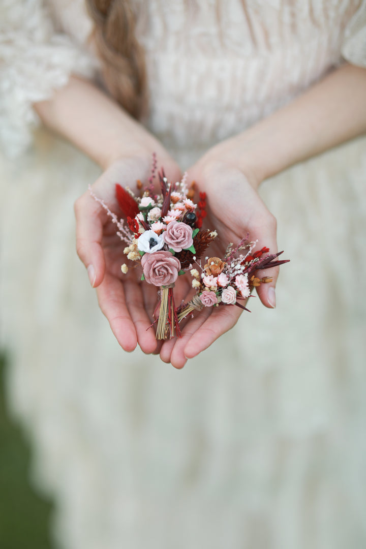 A person in a cream lace dress holds a small dried floral boutonniere in hands. The bouquet features dusty pink roses, red berries, and gold-toned accents.