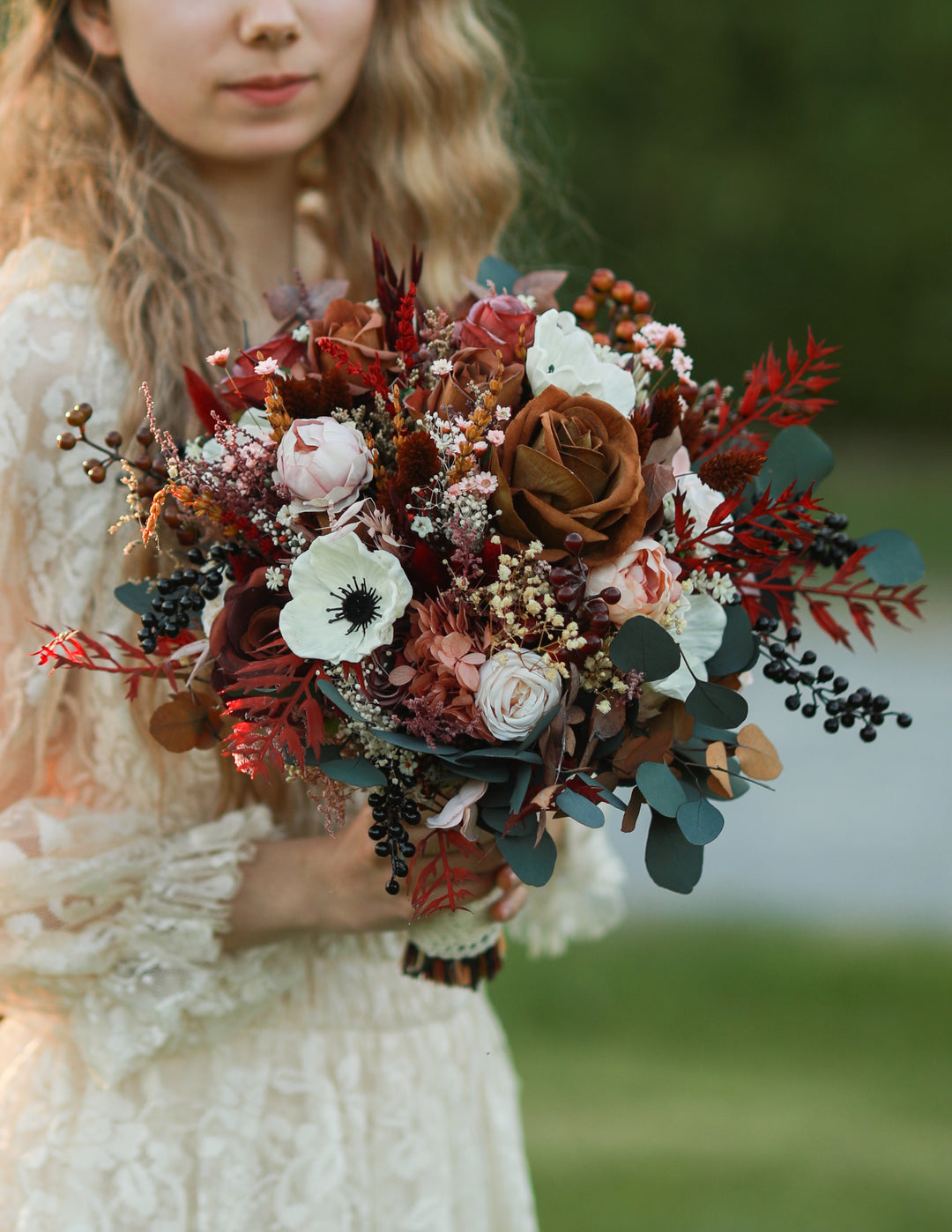 A woman in a cream lace dress holds a rustic autumn floral bouquet with red, brown, white, and green elements against a soft blurred outdoor background.