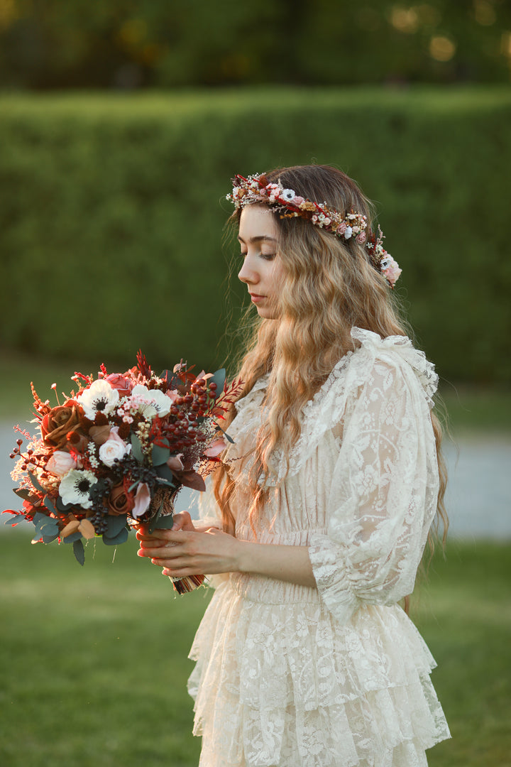 A woman in a bohemian-style cream lace dress and floral crown holds a rustic autumn bouquet in an outdoor garden setting.
