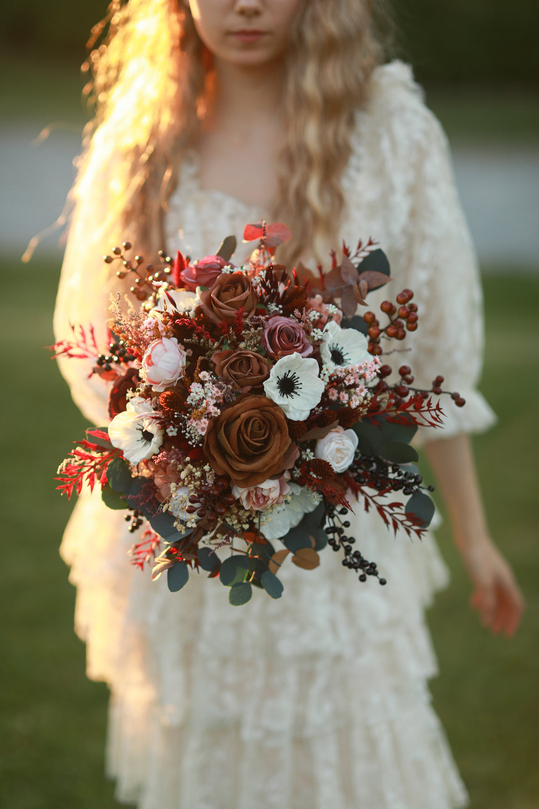 A woman in a white lace dress holding a rustic autumn floral bouquet with brown roses, white flowers, and muted greenery, photographed outdoors in warm golden light.