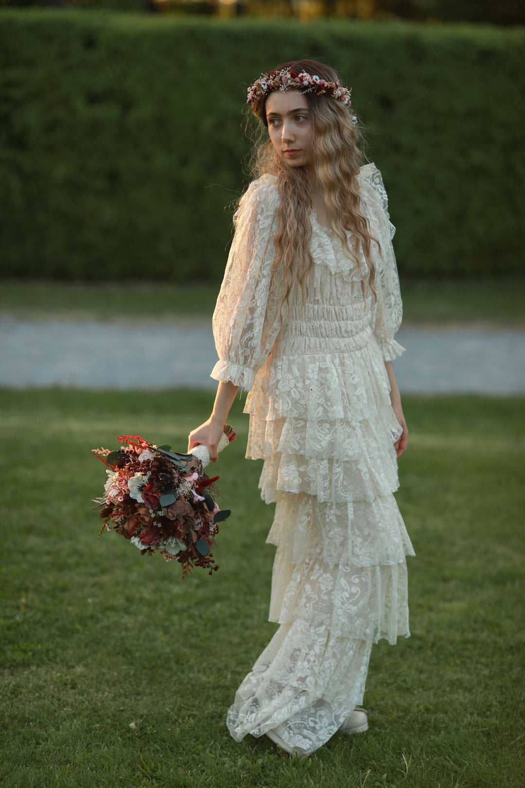 A woman standing on a grassy lawn wearing a long cream lace dress with ruffled sleeves and holding a bouquet of dark red and cream flowers. She has a floral crown on and is photographed outdoors against a green hedge.