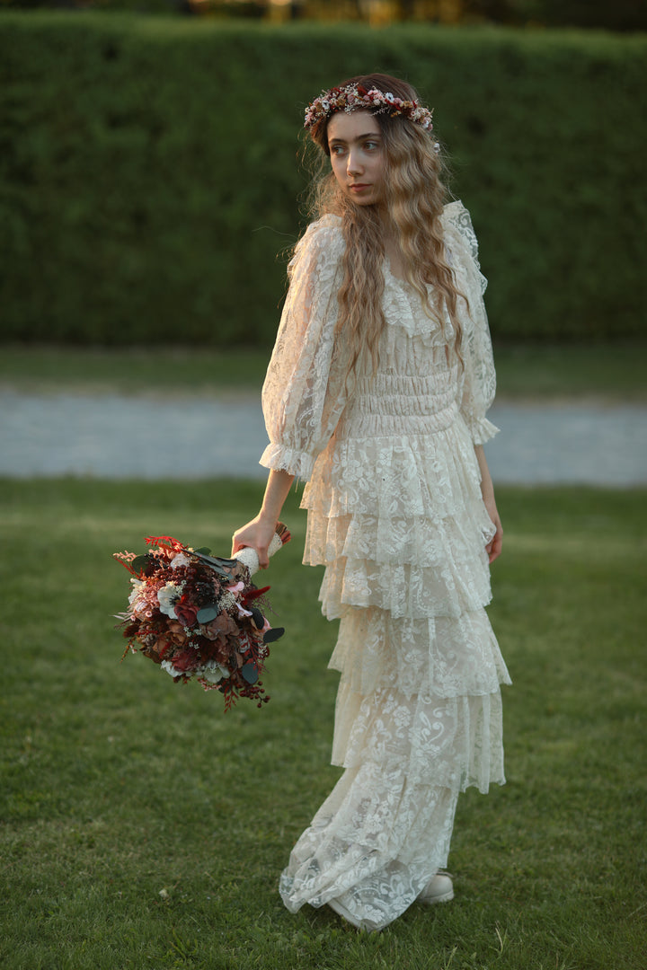 A woman standing on a grassy lawn wearing a long cream lace dress with ruffled sleeves and holding a bouquet of dark red and cream flowers. She has a floral crown on and is photographed outdoors against a green hedge.