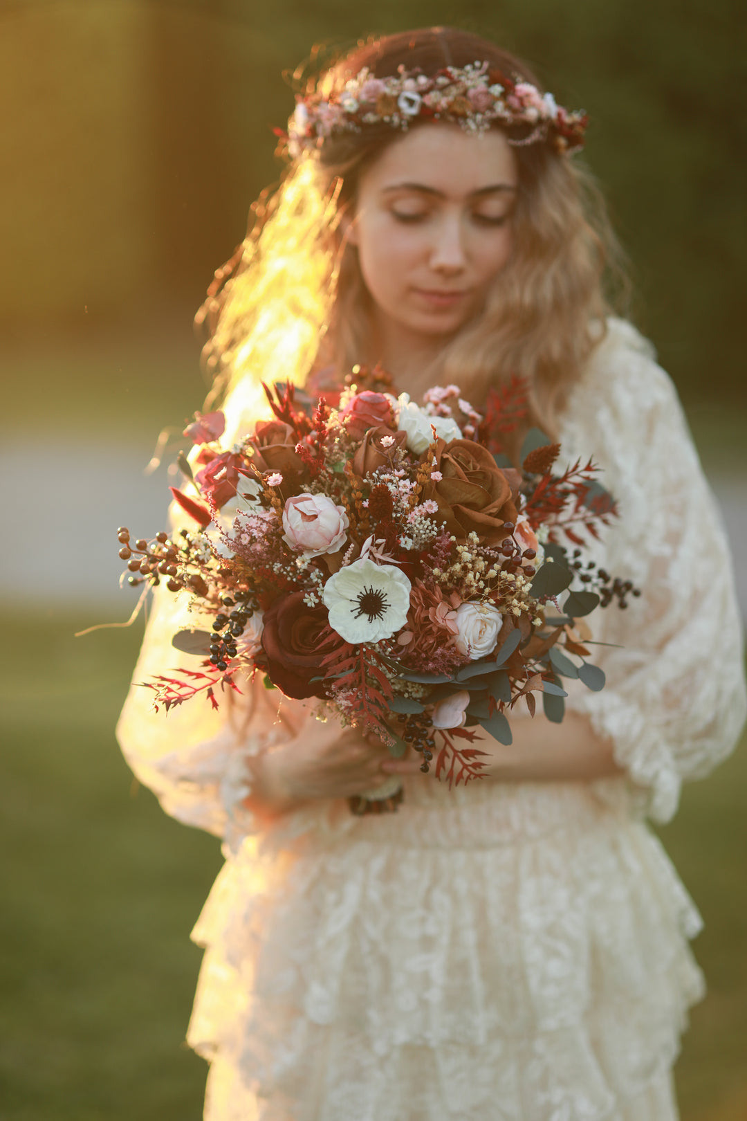 A woman outdoors wearing a cream lace dress and floral crown, holding a rustic bouquet of red, white, and green flowers. Warm golden sunlight backlights her from the side, with a softly blurred green background.