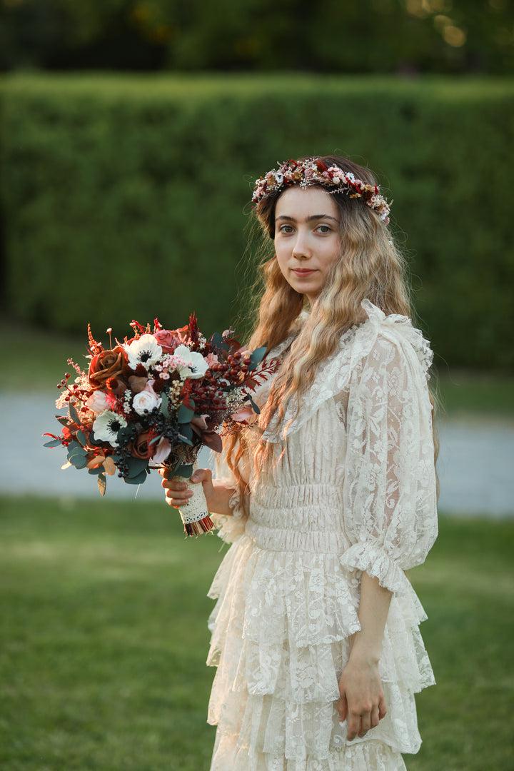 A woman standing outdoors in a grassy garden wearing a cream lace boho-style dress, a floral crown, and holding a rustic bouquet of red, white, and green flowers.