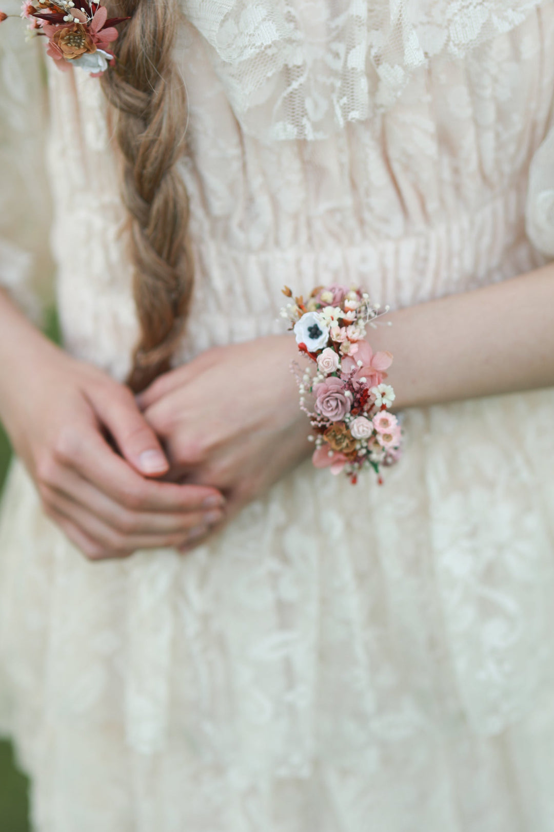A close-up lifestyle shot of a woman wearing a cream lace dress, featuring a delicate floral wrist corsage and a matching floral hair accessory.