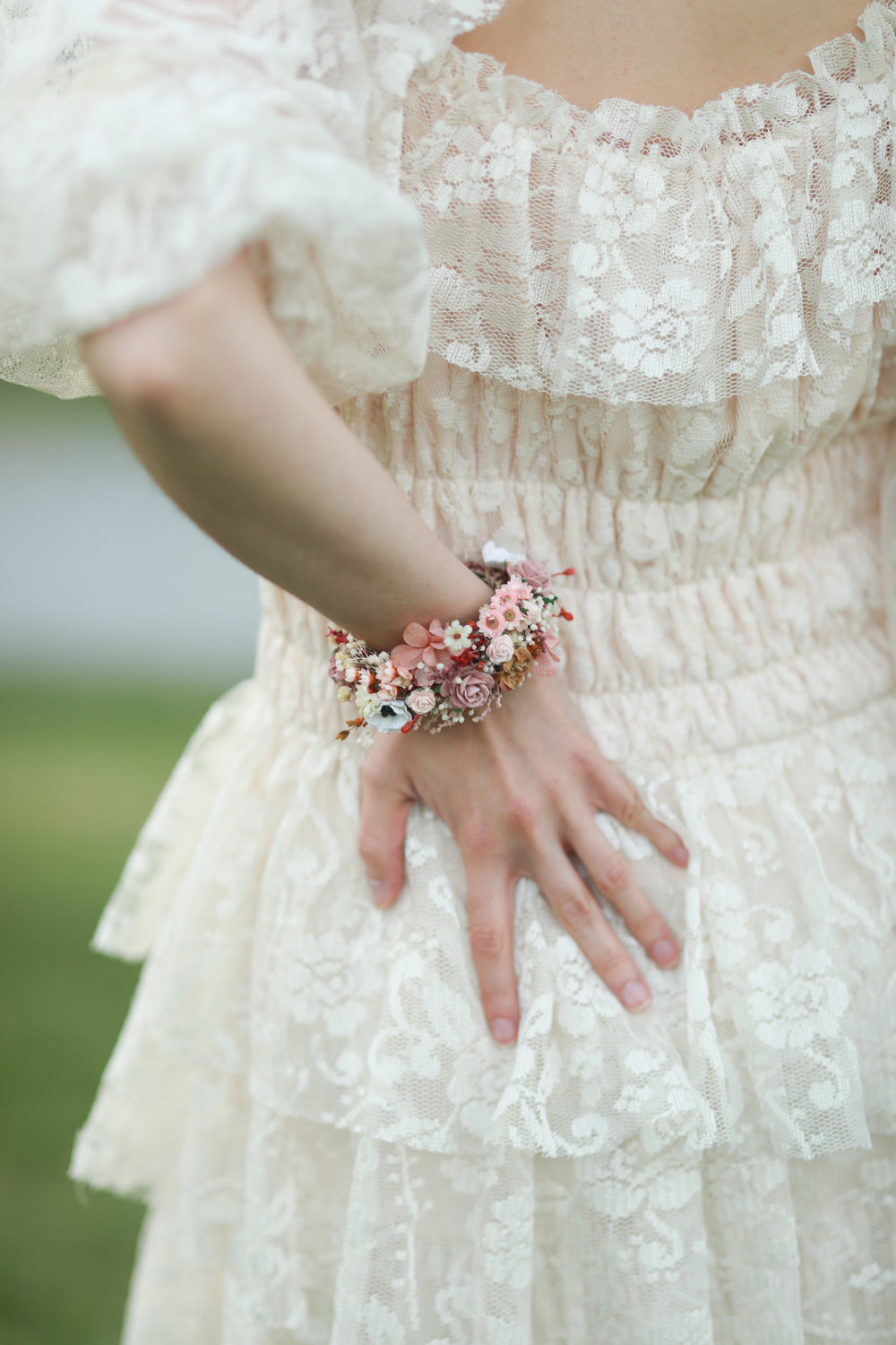 A close-up of a person wearing a cream lace dress and a floral wrist bracelet with pink, peach, and white flowers.