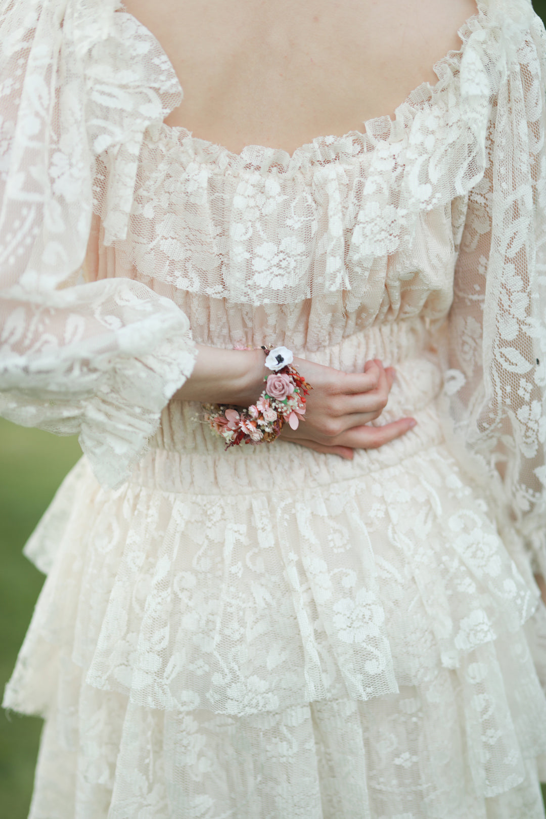 A close-up of a person wearing a cream lace dress with ruffled details and a floral bracelet on one wrist, photographed outdoors with a soft blurred green background.