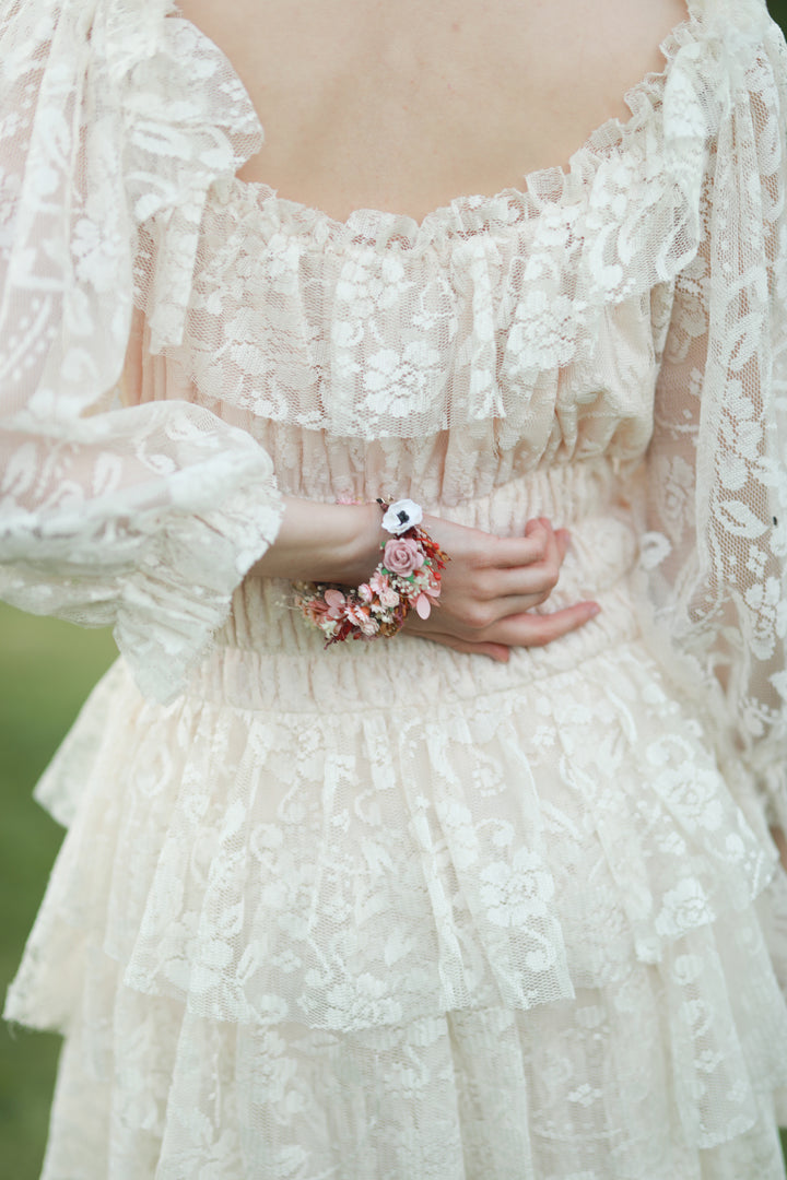 A close-up of a person wearing a cream lace dress with ruffled details and a floral bracelet on one wrist, photographed outdoors with a soft blurred green background.