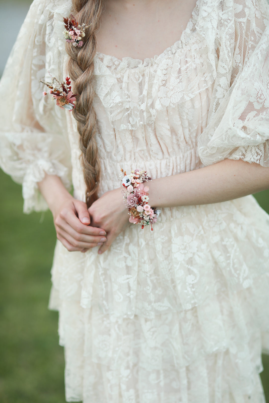 A close-up lifestyle shot of a woman wearing a cream lace boho dress, featuring a long side braid adorned with small pink and red floral hair clips and a matching floral wrist cuff bracelet.