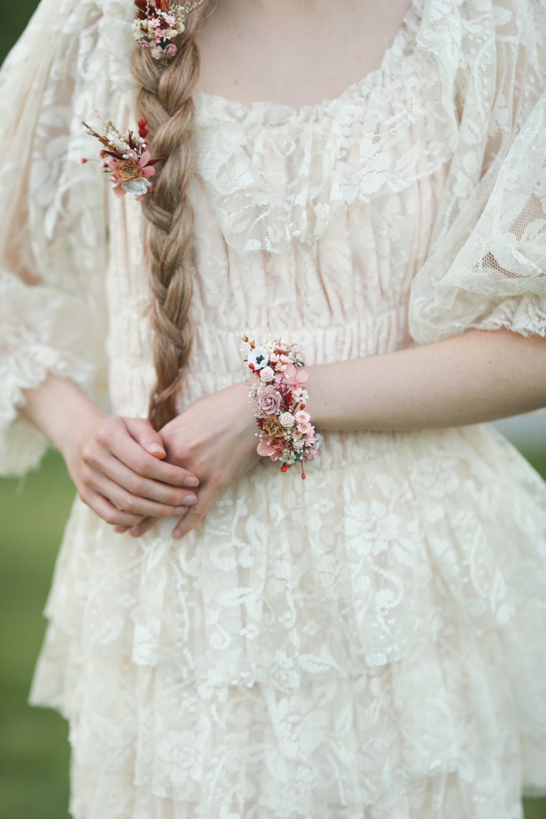 A close-up of a person wearing a cream lace dress outdoors, with a long braided ponytail and a floral wrist accessory visible.