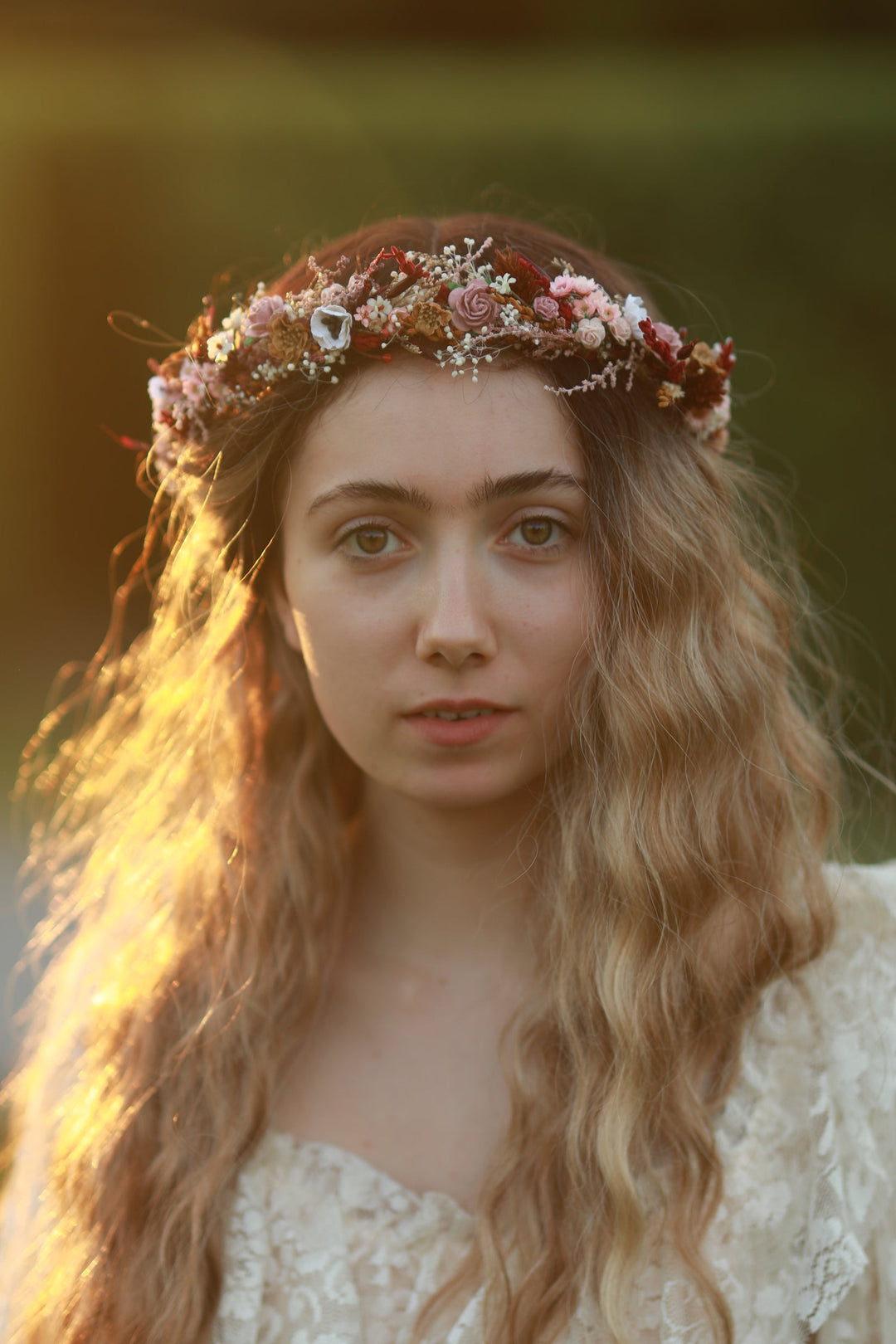 A woman with long wavy light brown hair wears a floral crown made of pink, red, and white flowers with small green leaves, set against a warm, blurred outdoor background.