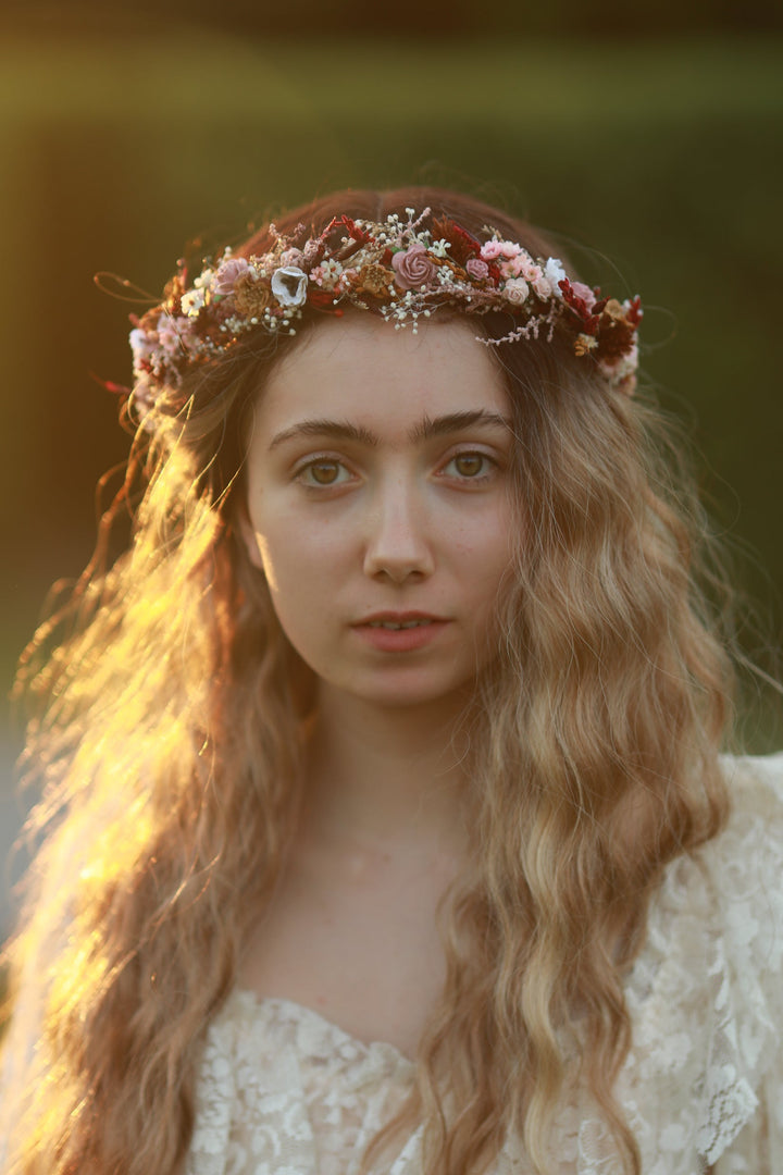 A woman with long wavy light brown hair wears a floral crown made of pink, red, and white flowers with small green leaves, set against a warm, blurred outdoor background.