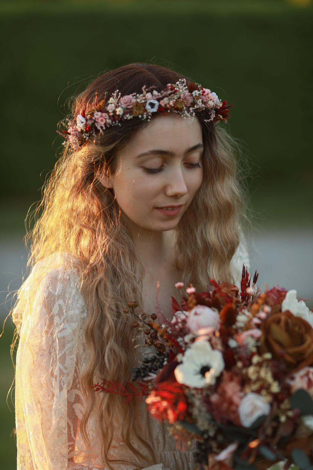 A woman outdoors wearing a floral crown and holding a bouquet of red, white, and brown flowers, with warm golden light on her hair and a soft blurred green background.
