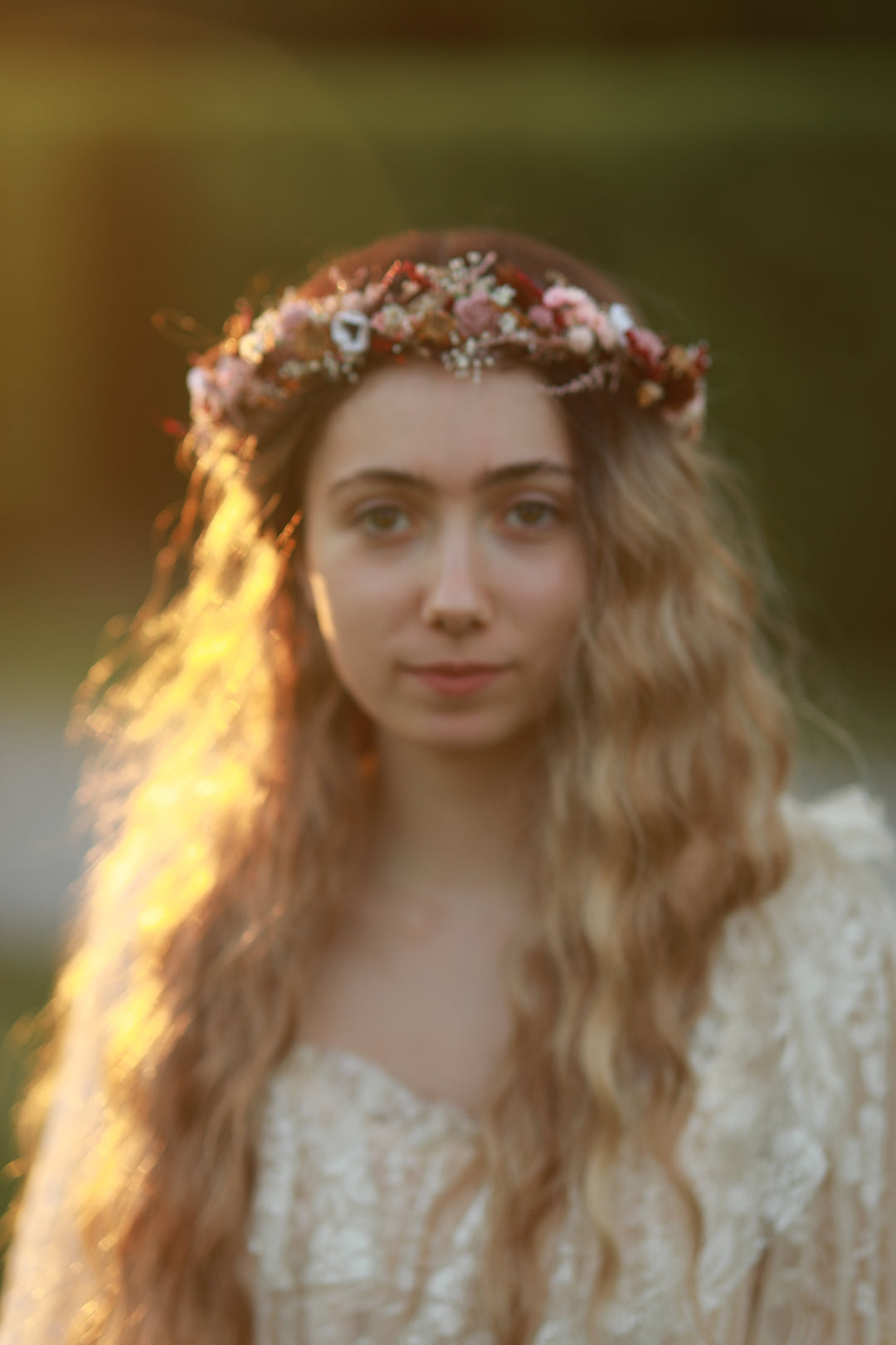 A woman with long wavy light brown hair wearing a floral crown of pink, white, and orange flowers, set against a warm, blurred outdoor background.