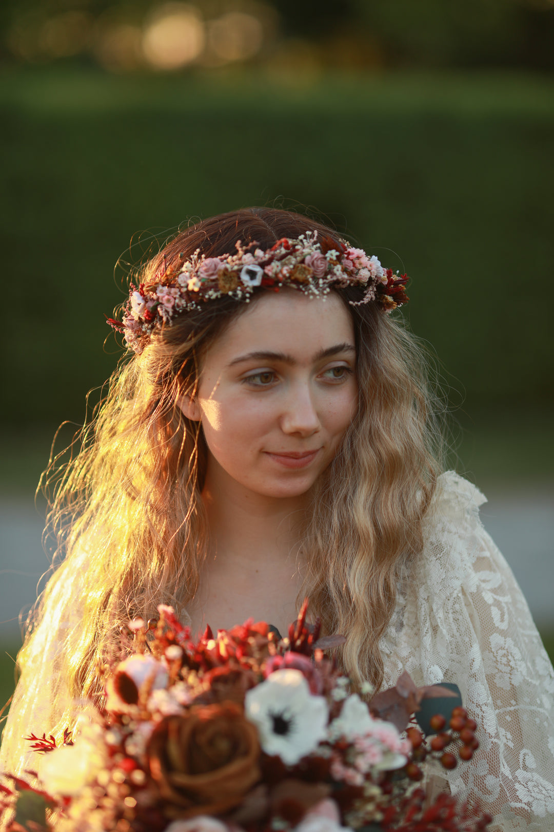 A woman outdoors wearing a floral crown and holding a bouquet of autumn-toned flowers, with warm golden light on her face and a blurred green background.