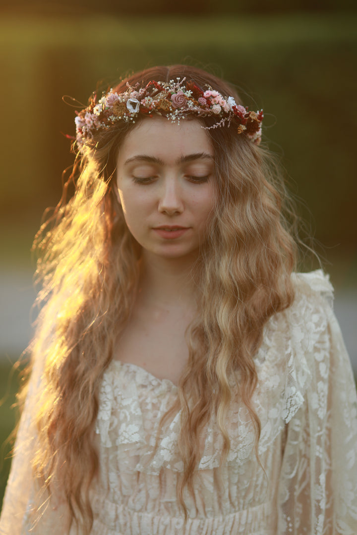 A woman with long wavy blonde hair wearing a floral crown and an off-white lace boho-style dress, captured in warm golden-hour sunlight against a blurred outdoor background.