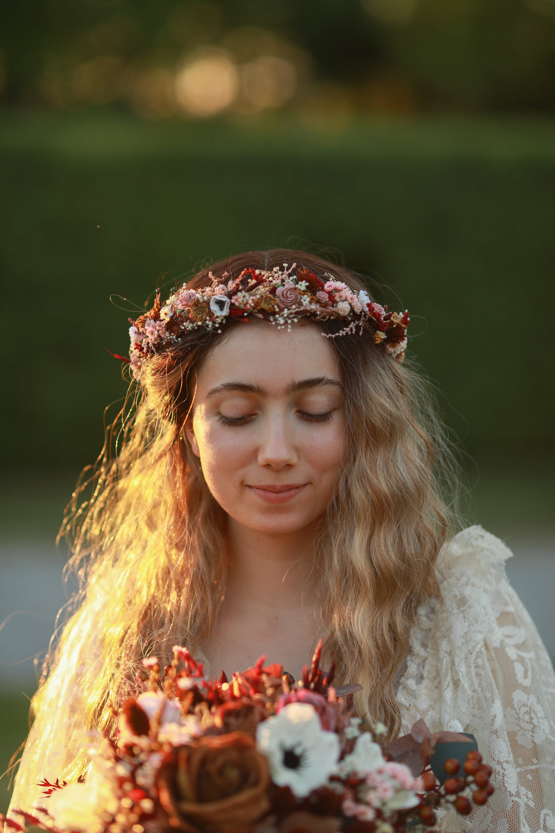 A woman outdoors wearing a floral crown and holding a bouquet of autumn-toned flowers, with warm golden light backlit against a blurred green background.