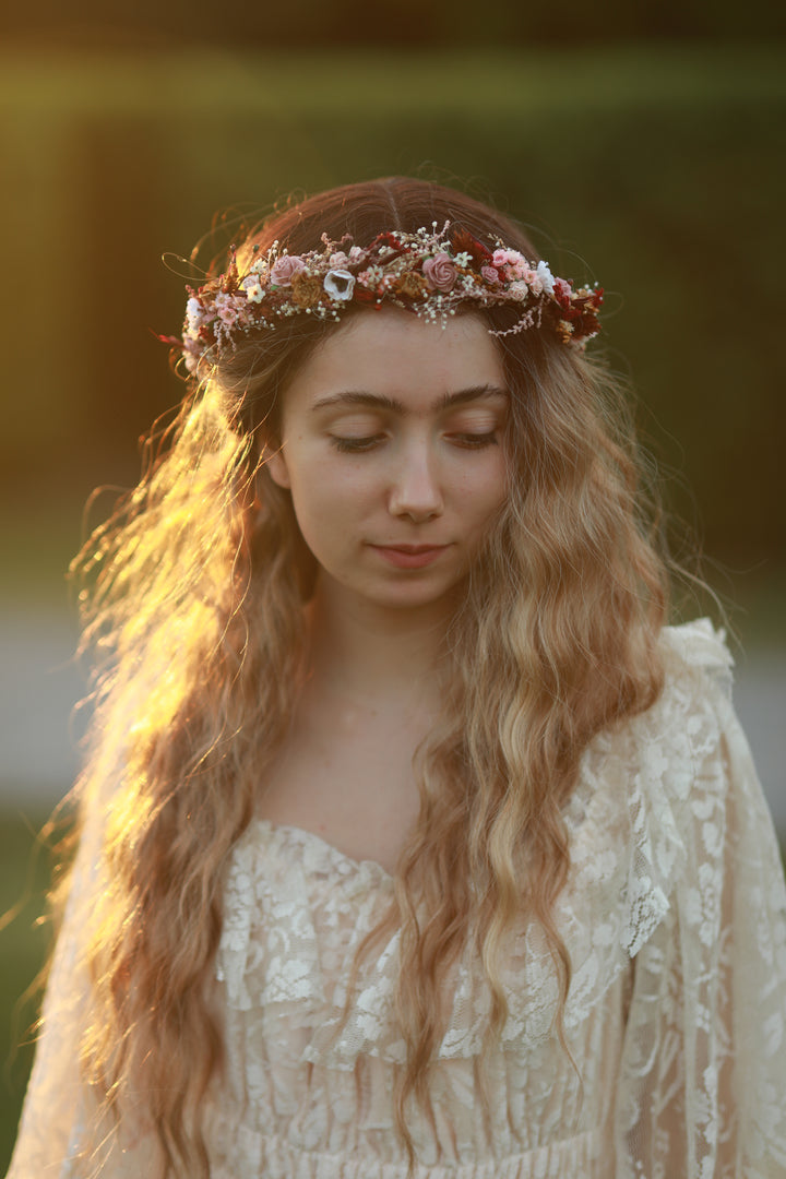 A woman outdoors wearing a bohemian-style cream lace dress and a floral crown of pink and white flowers, captured in warm golden hour sunlight.