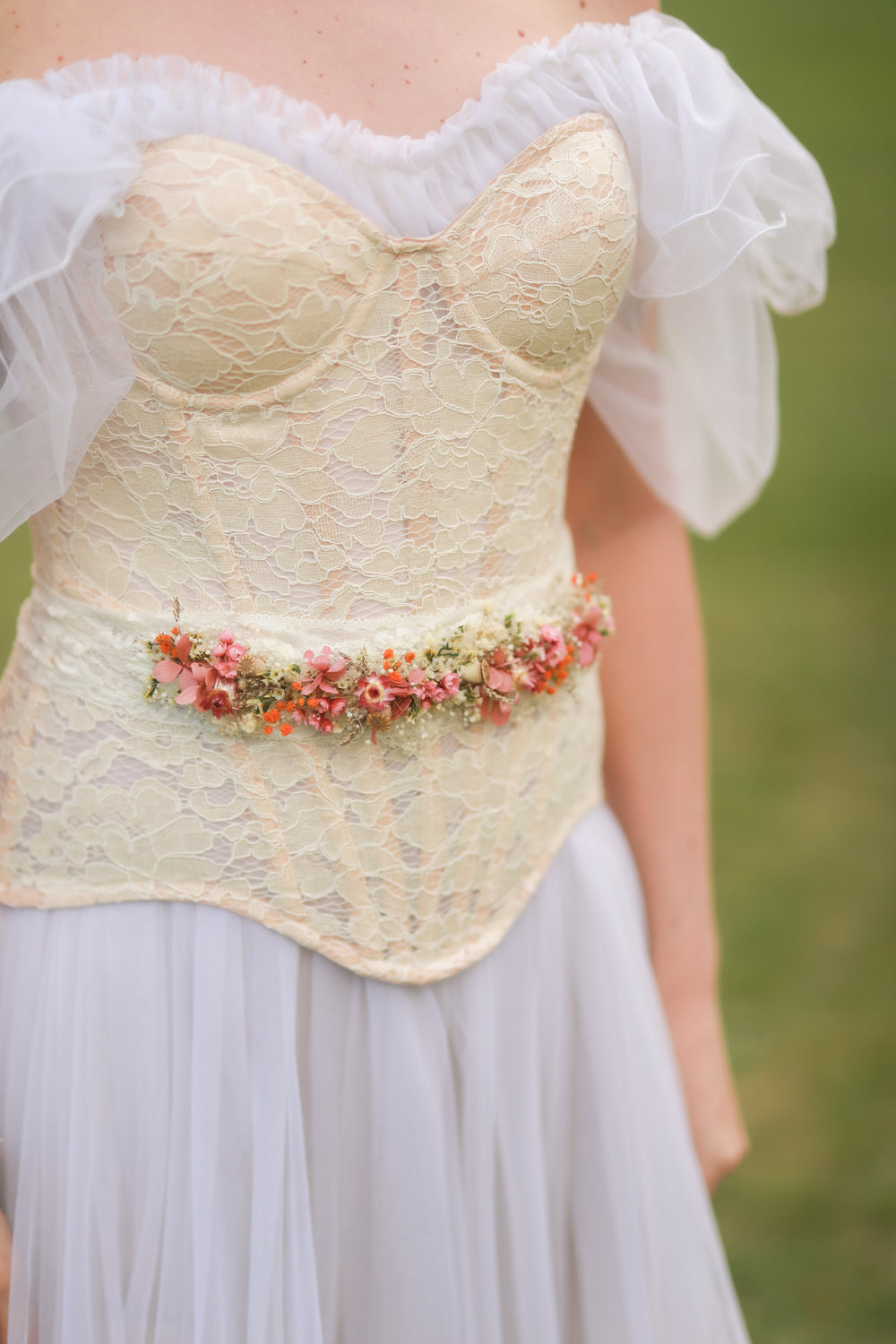 Close-up of a lace corset with floral belt worn by a person in a natural setting.