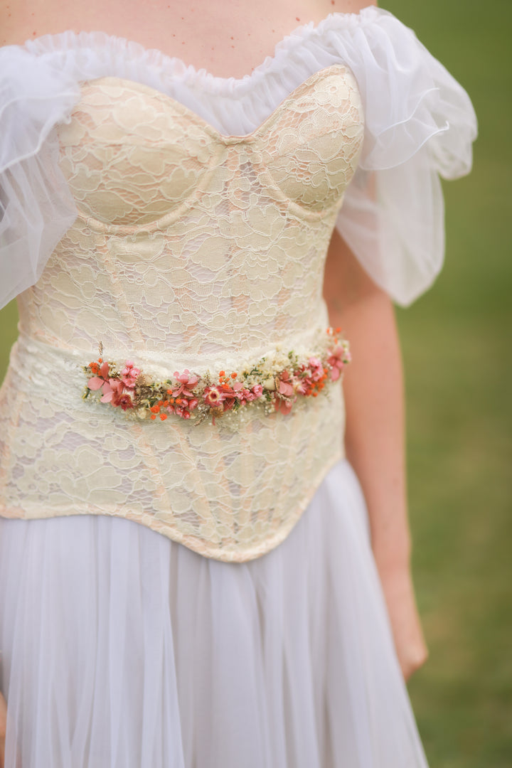 Close-up of a lace corset with floral belt worn by a person in a natural setting.