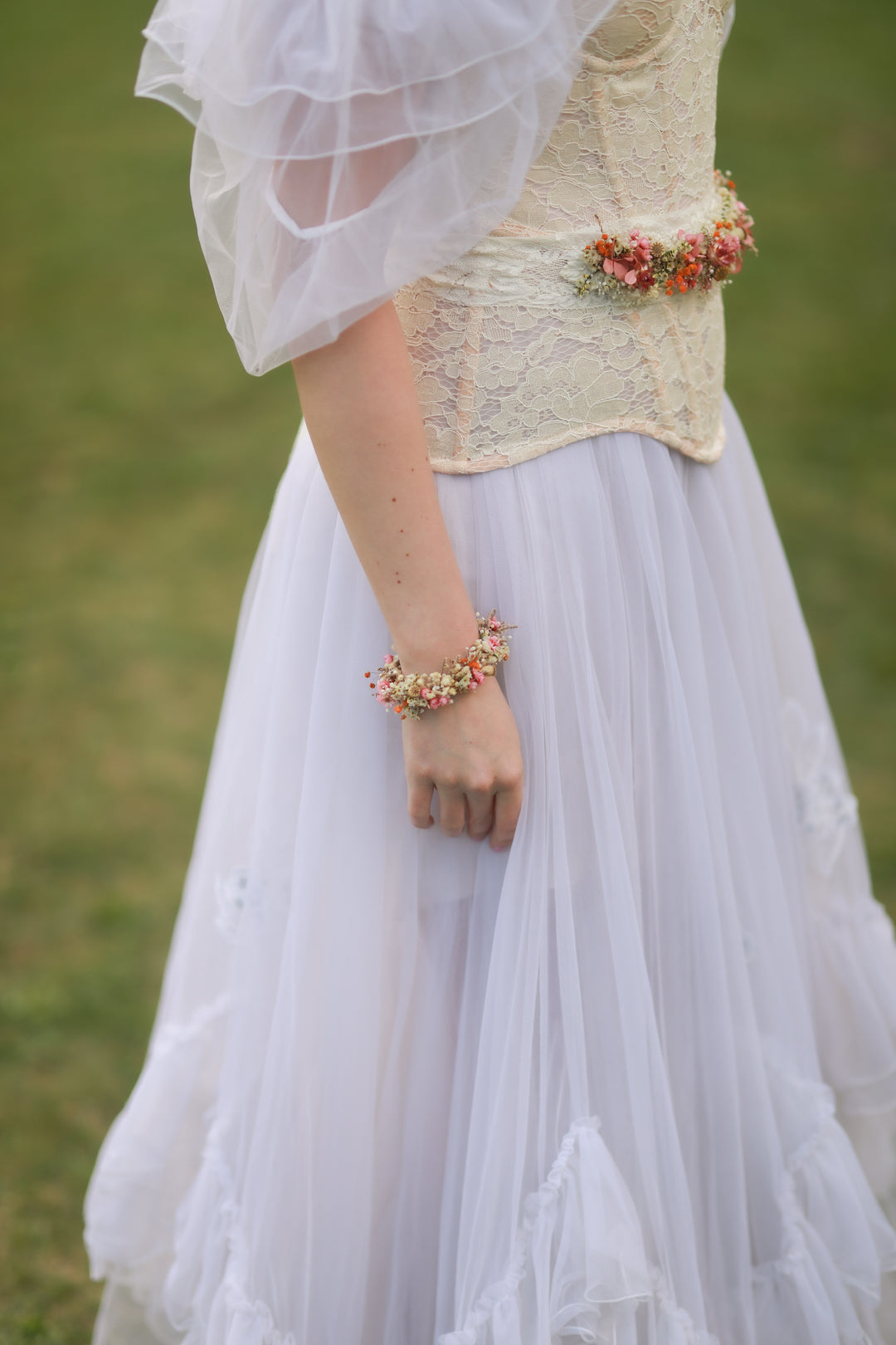 Person wearing a white lace top and light gray skirt with floral accessories against a blurred green background