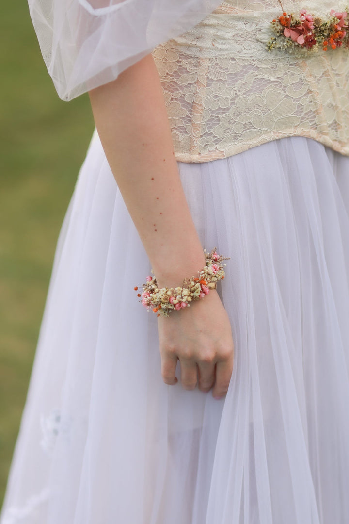 Person wearing a floral bracelet from dried flowers in pink, orange and cream tones  with a blurred natural background