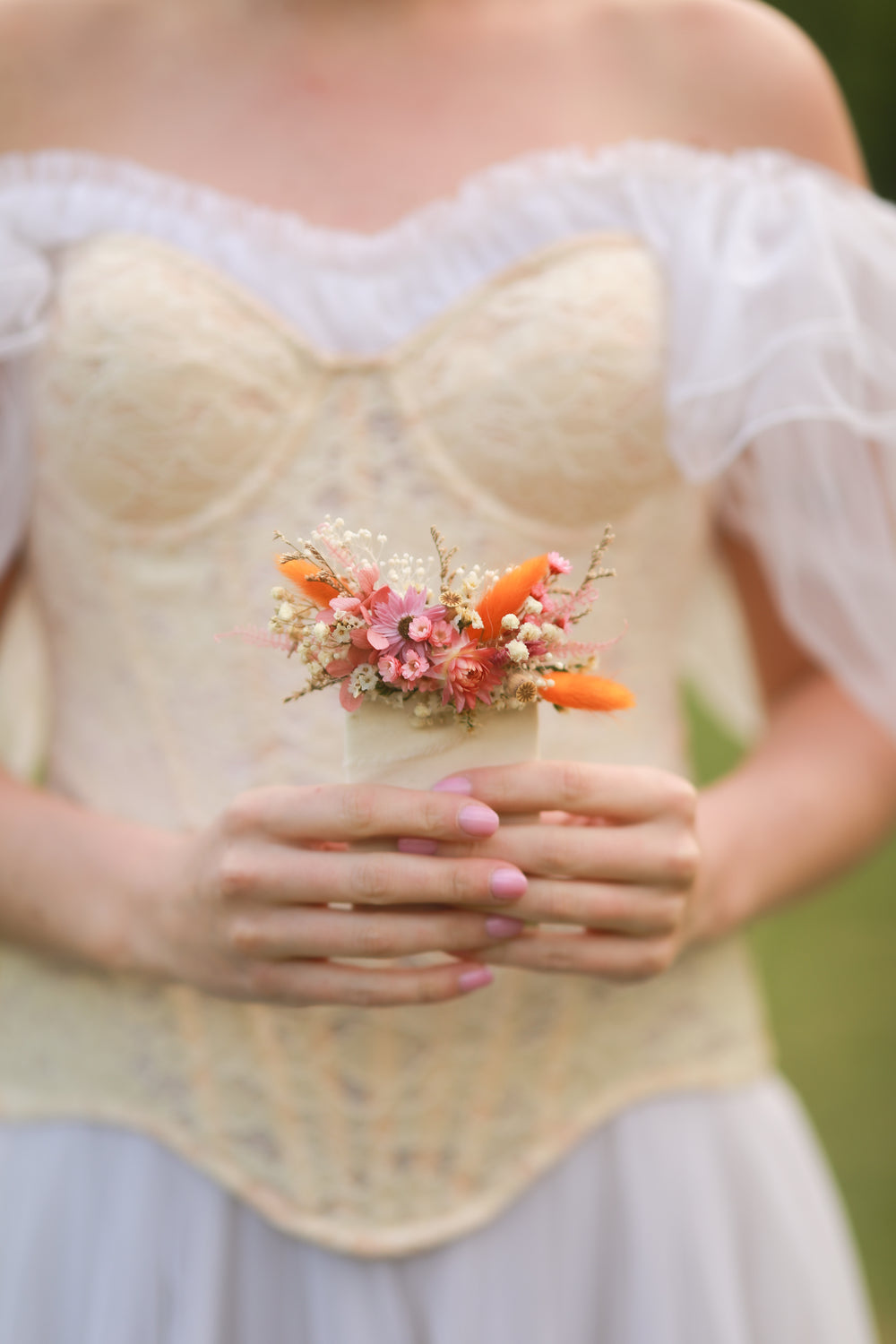 Person wearing a beige lace top with a floral boutonniere in pink, cream and orange tones held in front of their chest.