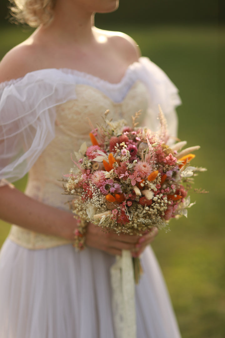 Person holding a bouquet of flowers with dried and prederved pink, orange and cream flowers in a natural setting