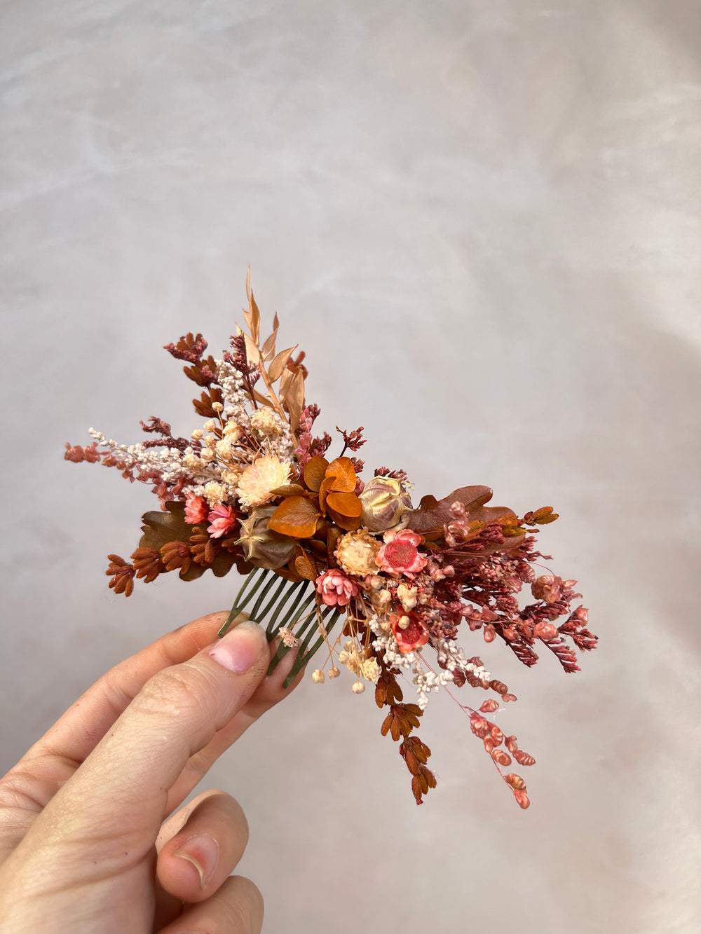 A hand holding a dried floral hair comb featuring autumnal shades of rust, terracotta, peach, and cream against a soft, textured light gray background.